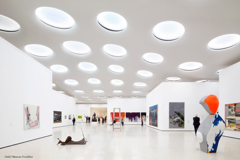Innenansicht des Städelmuseums mit runden Deckenleuchten. An den Wänden hängen verschiedene Kunstwerke, darunter Gemälde und Skulpturen. Eine Person steht im Raum.